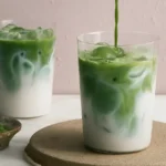 A glass of iced matcha latte with creamy green layers and ice cubes, served with a metal straw on a marble countertop